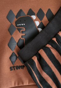 Sweat-shirt marron avec un motif de dinosaure noir et un motif en losanges, avec un bas en polaire rayé noir et marron. Texture douce.