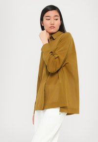 Camicetta marrone a maniche lunghe con bottoni e vestibilità comoda, dotata di colletto, fessure laterali e una sottile texture, abbinata a pantaloni bianchi.