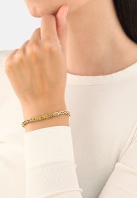 Złota bransoletka z ogniwami połączonymi ze sobą. Gładkie wykończenie i odbijająca światło powierzchnia. Noszona na nadgarstku nad żebrowanym, jasnym rękawem.