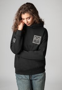 Zwarte hoodie met een voorzak en een gestructureerd patch met de tekst "BE THE ENERGY". Het heeft geribde manchetten en een losse pasvorm.