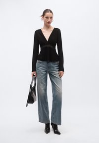 Bluză neagră cu mâneci lungi și detaliu din dantelă în talie; asortată cu blugi largi, decolorați, și tocuri negre cu vârf ascuțit; ține o geantă neagră.