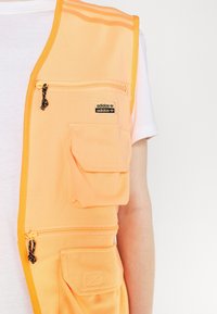 Orange tygväst med flera fickor, blixtlås och en liten svart Adidas-logotagg. Har kontrasterande gul kant och en slät textur.