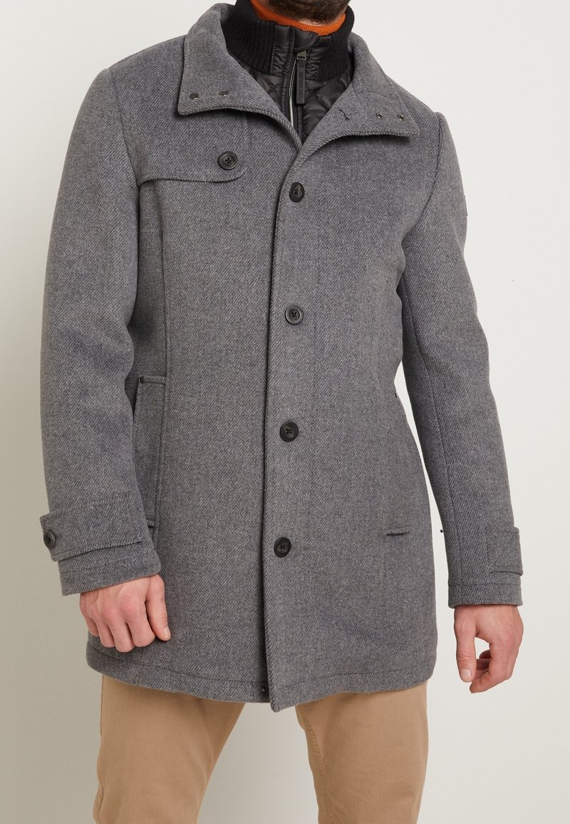 Homme portant un manteau en laine gris par-dessus une veste matelassée noire avec un pantalon marron clair, debout devant un fond clair uni.