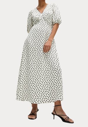 Kobieta w białej sukience midi z czarnym kwiatowym wzorem, bufiastymi rękawami i czarnych sandałach na obcasie z paskami, stojąca na jednolitym tle.