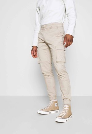 Mand iført beige cargobukser med elastiske manchetter og beige høje sneakers, stående mod en ensfarvet hvid baggrund.
