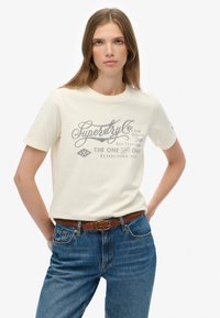 Kvinde med langt brunt hår iført en beige Superdry Co. T-shirt, blå jeans og et fletet brunt bælte, stående med hænderne bag ryggen.