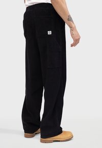 Sorte kordfløyels vide bukser med to store sidelommer og elastisk midje, sammen med brune clogs på en ensfarget bakgrunn.