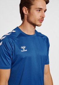 Mėlynas sportinis marškinėliai, pagaminti iš lygaus audinio, su baltais dryžiais ant pečių ir spausdintu logotipu ant krūtinės. Trumpi rankovės.