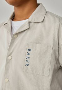 Button-up-Hemd in hellem Beige mit subtiler gestreifter Textur. Verfügt über eine Brusttasche mit dem Wort "BAKER" in navy Buchstaben.