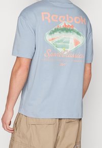 Světle modré bavlněné tričko se grafickým potiskem na zadní straně, s textem a diamantovým designem znázorňujícím sportovní hřiště.