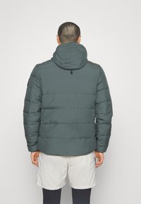 Dunkelgrüne Pufferjacke mit Kapuze, gestepptem Design und elastischen Bündchen. Sichtbare Nähte und ein kleines Logo auf der Schulter.