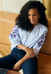 Blouse rayée bleu clair et blanche avec broderie florale violette sur les manches. Présente un col rond et une fermeture à boutons.