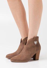 Brune ruskindsstøvler til anklen med spids tå, hælafslutning og dekorativ løkke. Har en struktureret, stablet træsålshæl.