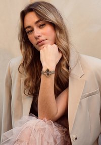 Bracelet en or avec un design floral et une sangle grise. Le modèle porte un blazer beige avec une jupe rose à volants, sur un fond neutre.