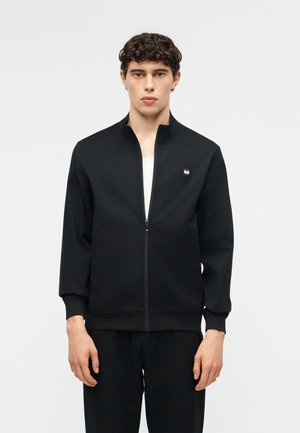 Ung mand med krøllet hår iført en sort lynlåstrøje over en hvid skjorte, stående mod en ensartet lys baggrund.