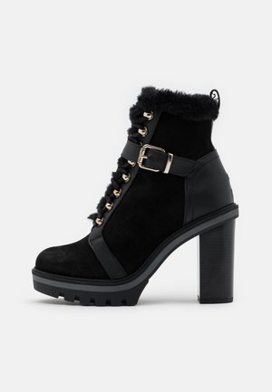 Botte noire à la cheville en daim et cuir, avec un bord en fourrure, des œillets dorés, une sangle avec boucle, un talon épais et une semelle texturée.