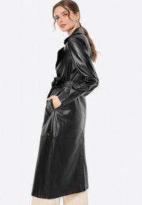 Cappotto trench in pelle nera con vita cinturata, maniche lunghe e tasche. La texture liscia e il design elegante si estendono fino a un'altezza media del polpaccio.