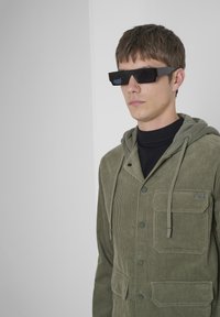 Chaqueta de pana verde oliva con capucha, bolsillos frontales y cierre de botón. Llevada sobre un cuello de tortuga negro y combinada con gafas de sol negras angulares.