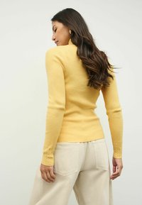 Frau mit langen dunklen Haaren, die einen gelben gerippten Rollkragenpullover und beige Hose trägt, leicht seitlich vor einem schlichten Hintergrund positioniert.