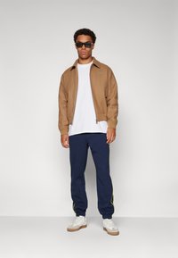 Casaco castanho com fecho éclair sobre uma t-shirt branca, calças azul-marinho com riscas laterais verdes e ténis brancos. Óculos de sol modernos completam o visual.