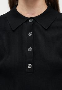 Κοντινό πλάνο μαύρου πλεκτού πόλο με ραβδώσεις και ασημένια κουμπιά με λογότυπο στο εμπρόσθιο μέρος κάτω από γιακά με μυτερή άκρη.