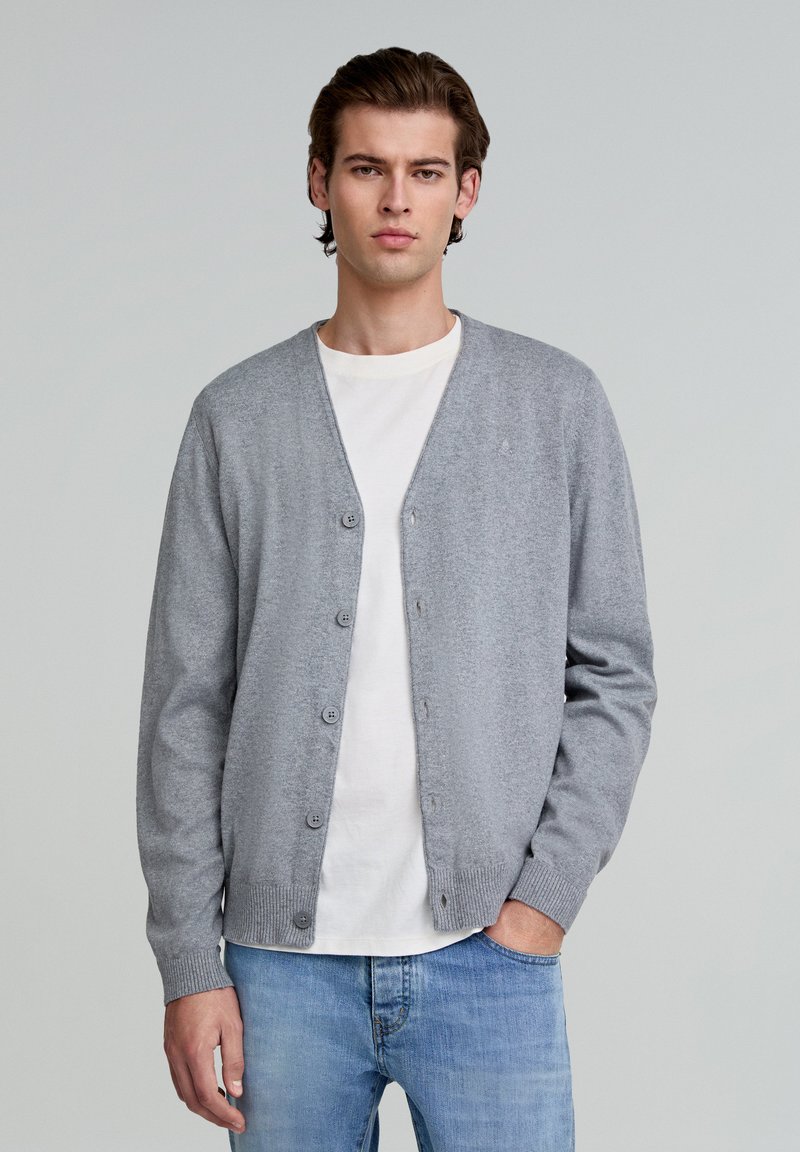 Grå cardigan lavet af blødt strikstof med V-hals, knaplukning, ribbedeærmer og en ensfarvet struktur. Båret over en hvid t-shirt.