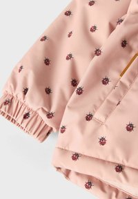 Gaiši rozā audums ar izdrukātām sarkanu un melnu vabolu formām, ar elastīgu aproci un redzamu šuvju, kā arī zelta brūnu etiķetes detaļu.