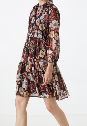 Femme portant une robe mi-longue à manches longues, avec une base noire et un grand imprimé floral aux tons rouges, beige et blancs.