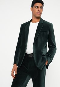 Sötétzöld bársonykabát, slim fit szabással, hajtókás gallérral, egyetlen mellzsebbel és oldalzsebekkel, fehér pólóval és fekete övvel párosítva.