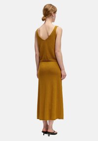 Senfgelbes, geripptes Strickkleid mit tiefem V-Ausschnitt und taillierter Passform, das bis zur Mitte der Wade fließt, kombiniert mit schwarzen Sandalen mit Absatz.