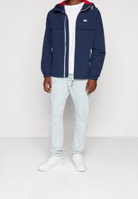 Námořnicky modrá lehká bunda s červenou podšívkou kapuce, předním zipem a elastickými manžetami, nošená přes bílé tričko a světle modré džíny, doplněná bílými teniskami.