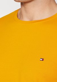 Närbild av en person som bär en senapsgul crewneck-tröja med en liten rektangulär röd, vit och blå logotyp på bröstet.