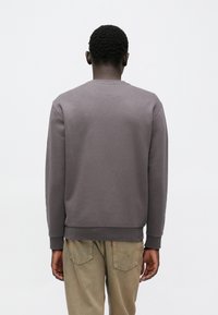 Personne aux cheveux courts et bouclés, portant un sweat-shirt gris foncé uni et un jean marron clair, vue de dos sur un fond blanc.