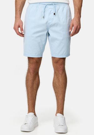 Shorts à cordon bleu clair pour homme portés avec des baskets blanches et une chemise blanche, debout devant un fond uni.