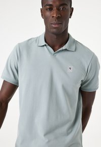 Polo de color azul claro hecho de una tela suave, con una botonadura de tres botones y un pequeño parche con el logo en el lado izquierdo del pecho. Manga corta.