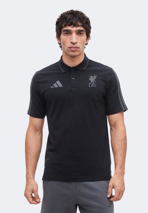 Homme portant un polo Adidas Liverpool FC noir avec logo sur la poitrine et trois bandes sur les manches, debout devant un fond uni.