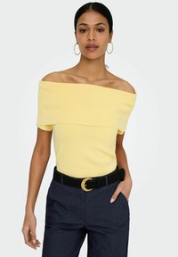Femme portant un haut jaune à épaules dénudées, un pantalon foncé avec une ceinture noire ornée d'une boucle dorée, et de grandes boucles d'oreilles créoles.