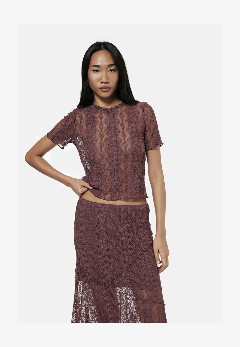 Woman with long dark hair wears matching sheer lace purple top and skirt, standing with one hand on hip against plain white background.