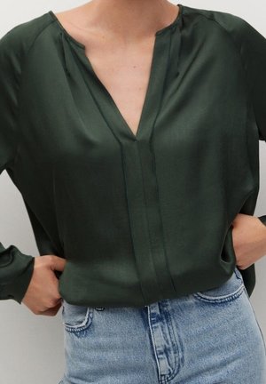 Osoba w ciemnozielonej bluzce z dekoltem w serek i długimi rękawami, włożonej do jasnoniebieskich wysokich dżinsów, ręce spoczywają blisko kieszeni.