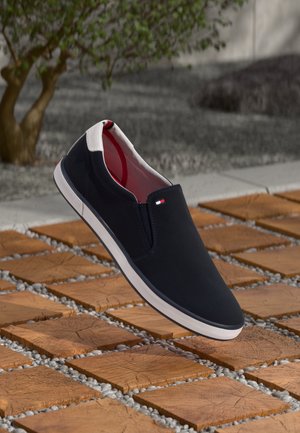 Sort slip-on sneaker med hvid sål og hælflig, lille rød-hvid-blåt logo, vist over træfliser med grus og en plante i baggrunden.