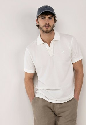 Hombre con una camisa polo blanca de manga corta, pantalones beige y una gorra de béisbol gris oscuro, de pie con las manos en los bolsillos.