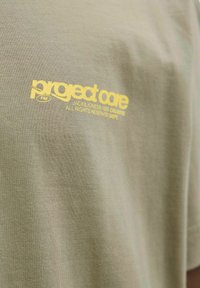 Olivenfarvet bomulds T-shirt med korte ærmer, der har et gult trykt logo nær halsudskæringen, som siger "project core".