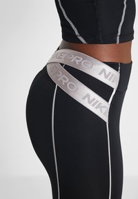Czarne legginsy sportowe mają szeroki, skrzyżowany szary pas z napisem "NIKE PRO". Projekt obejmuje białe szwy i dopasowany krój.