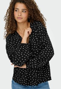 Zwarte blouse met wit bloemenpatroon, v-hals, lange pofmouwen, knoopsluiting aan de voorkant, gestructureerde stof. Draagt met lichtblauwe denim jeans.