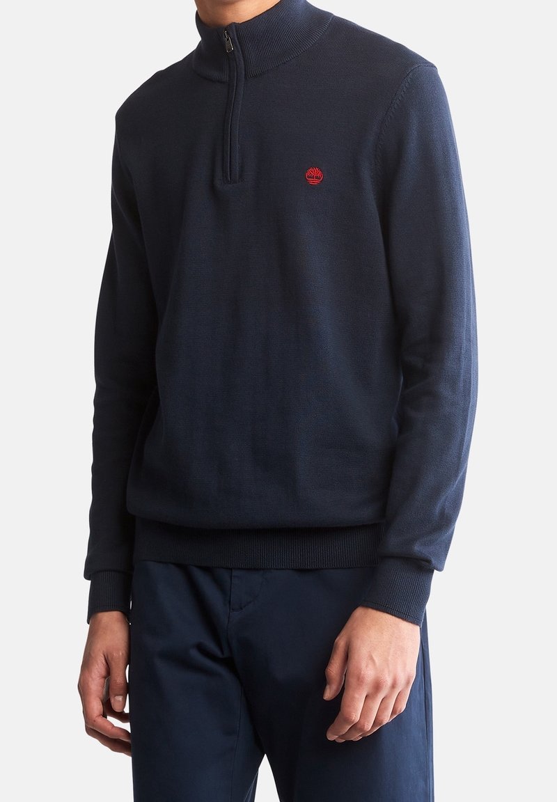 Suéter de punto azul marino con cuello con cremallera, puños y dobladillo acanalados, que presenta un pequeño logo rojo en el pecho. Textura suave, ajuste informal.