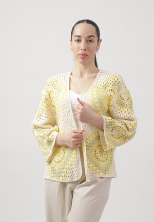 Donna con capelli scuri raccolti indossa una maglietta bianca e pantaloni beige con un cardigan lavorato a uncinetto, ampio, giallo e lavanda.