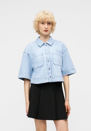 Young person with short curly blonde hair wearing a light blue cropped denim jacket and a black pleated skirt, standing against a plain background.