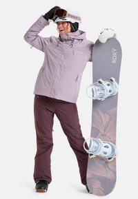 Snowboarder in a light lavender jacket and maroon pants holds a gray snowboard with floral patterns and white bindings, wearing a helmet and gloves.