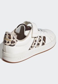White athletic shoe with a textured synthetic upper, leopard print accents, adjustable strap, and rubber sole. Features Adidas branding.