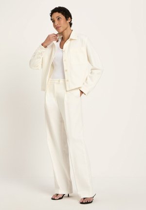 Frau, die eine cremeweiße, geknöpfte Jacke, passende weit geschnittene Hosen, ein weißes Tanktop und schwarze Sandalen mit Absatz trägt und vor einem schlichten Hintergrund steht.